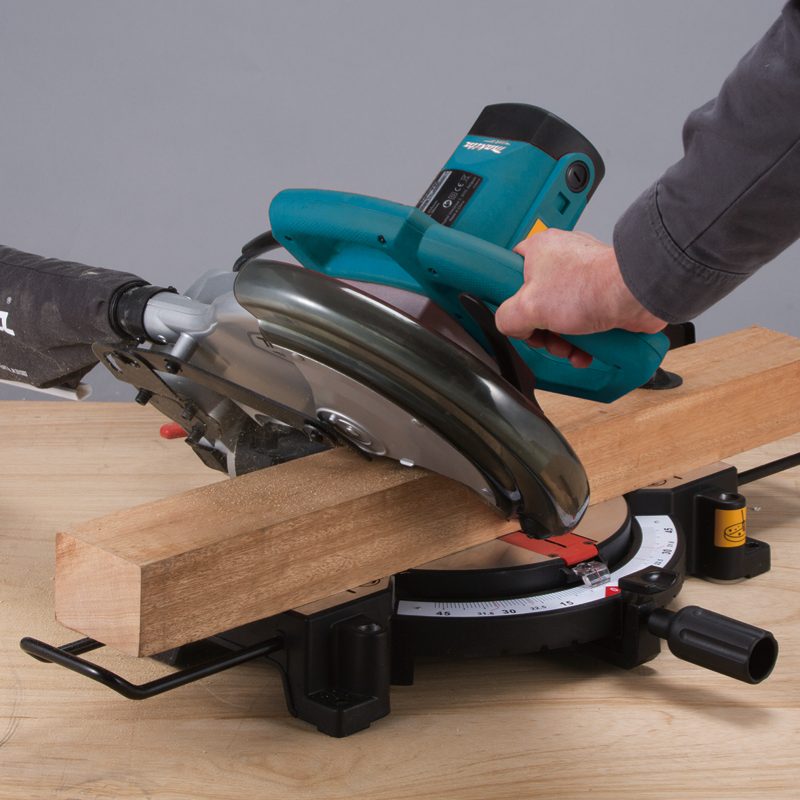 Makita M2300B 255mm compound miter saw cutting a timber board with a vacuum cleaner connected for dust extraction