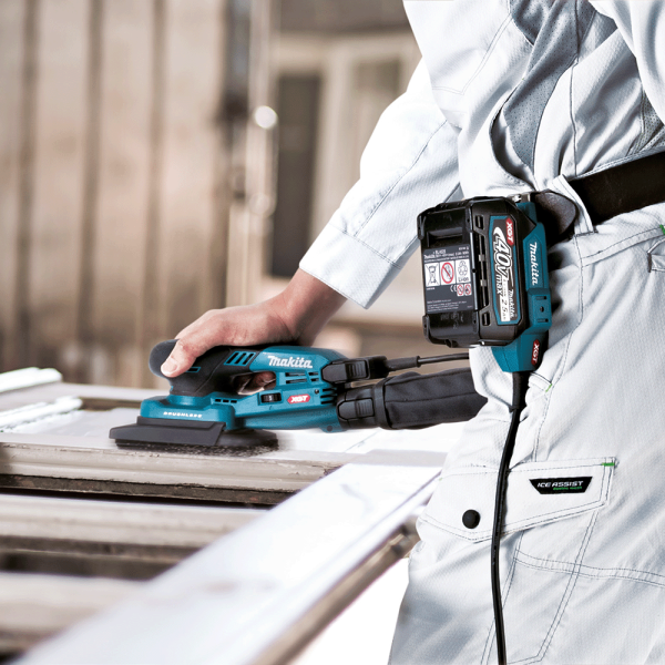 Worker sanding a door frame with the Makita BO005CGZ 40V cordless orbital sander, battery carried on the belt via the adapter