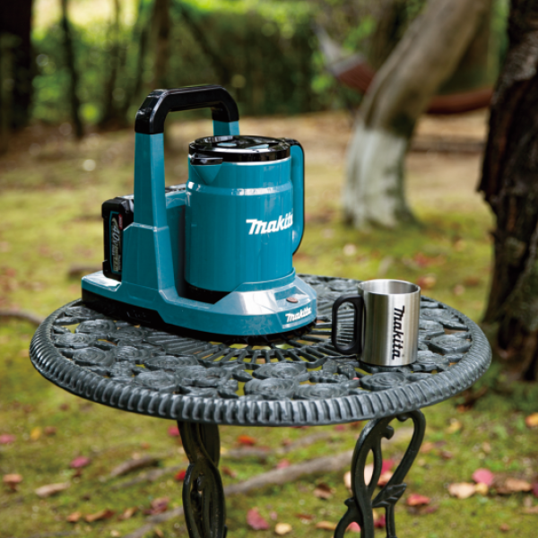 Makita KT001GZ 40V cordless kettle on its power supply stand outdoors, with a stainless steel mug beside it