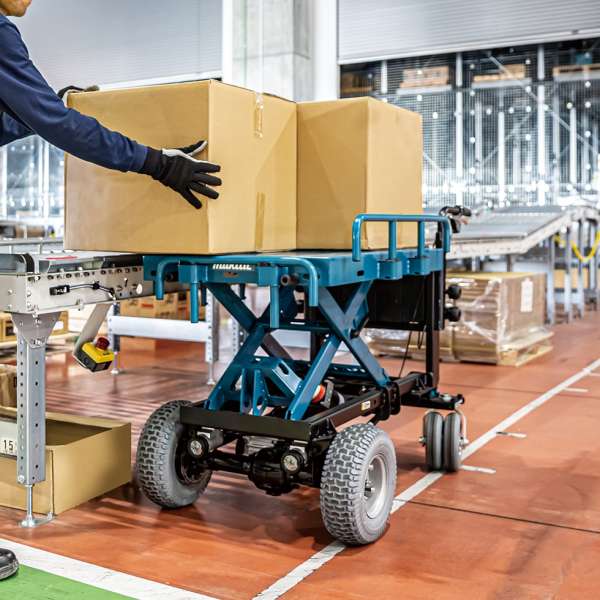 Operator loading a carton onto the raised electric lift deck of a Makita DCU601Z wheelbarrow in a warehouse