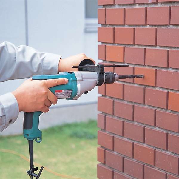 Operator drilling into a red brick wall with a Makita HP2050 hammer drill fitted with side grip and depth gauge