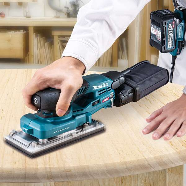 Makita BO007CGZ 40V cordless orbital sander sanding a wooden board with the dust bag fitted and the battery worn separately on an adapter