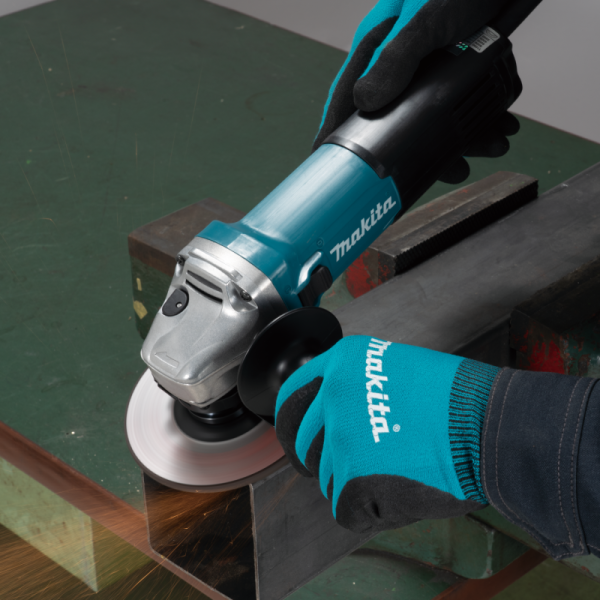 Gloved operator using the Makita GA5100 angle grinder on a metal workpiece at a workbench