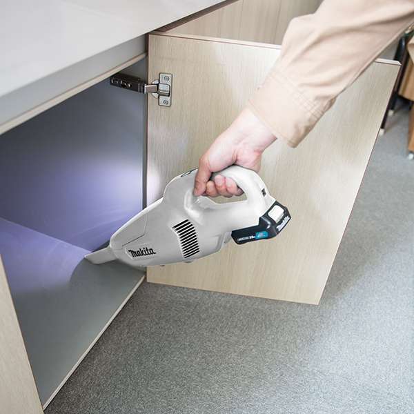 Makita CL107FDWAW 12V cordless handheld vacuum cleaner in white, used to clean under a kitchen cabinet